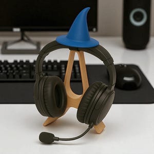 以下が含まれることがあります： 黒いオーバーイヤーヘッドホンとマイクが木製のスタンドに置かれています。小さな青い魔法使いの帽子が上に乗っています。スタンドは白い表面にあり、背景にはコンピューターモニターとキーボードがあります。