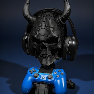 以下が含まれることがあります： 角のある黒い頭蓋骨型のヘッドフォンスタンド。黒いヘッドフォンがかけられています。青いゲームコントローラーが頭蓋骨の下の黒いベースに置かれています。スタンドは濃い青色の表面にあります。