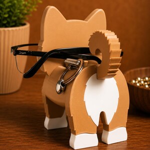 May include: A wooden corgi-shaped glasses holder in shades of brown and white, with black-framed glasses resting on it. The holder features a safety pin detail and a fluffy tail. The design is whimsical and functional, perfect for desk organization.