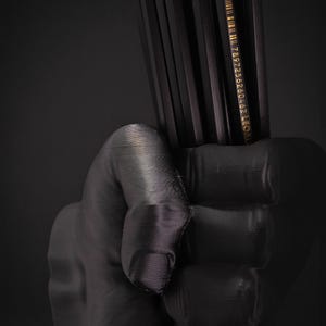 May include: A black, 3D-printed hand holding a bundle of black pencils. The pencils have gold text and a barcode. The image is set against a dark background, creating a dramatic contrast.