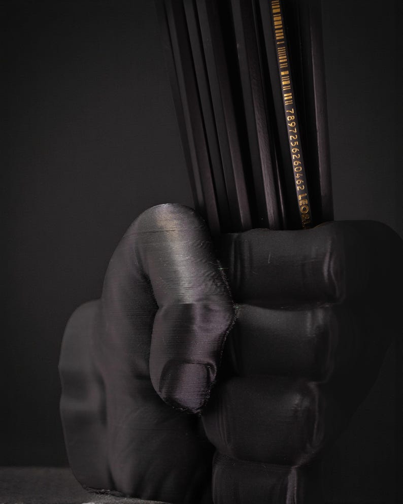May include: A black, 3D-printed hand holding a bundle of black pencils. The pencils have gold text and a barcode. The image is set against a dark background, creating a dramatic contrast.