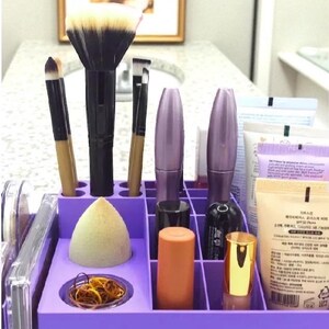 May include: A purple makeup organizer filled with various beauty products. The organizer holds makeup brushes, mascara tubes, a makeup sponge, lipsticks, and other cosmetic items. The background includes a bathroom sink and a framed picture.