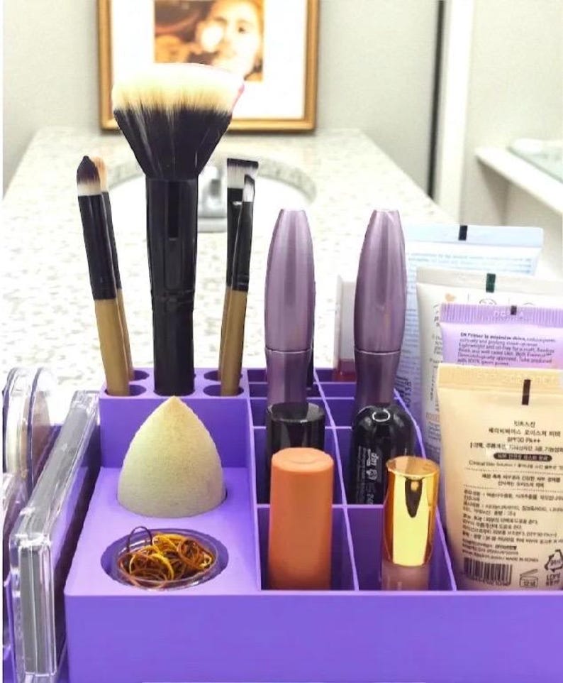 May include: A purple makeup organizer filled with various beauty products. The organizer holds makeup brushes, mascara tubes, a makeup sponge, lipsticks, and other cosmetic items. The background includes a bathroom sink and a framed picture.