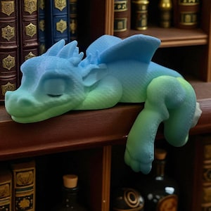 May include: A sleeping dragon figurine in shades of blue and green rests on a wooden shelf. The fantasy creature has closed eyes, wings, and detailed scales. The figurine is a decorative item.