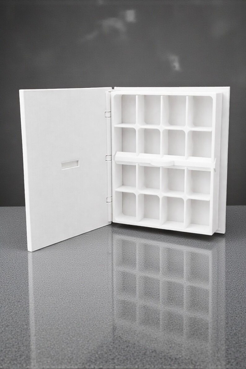 Peut inclure: Un organisateur blanc ouvert pour bijoux ou petits objets, avec de multiples compartiments carr&eacute;s. L'organisateur a une porte &agrave; charni&egrave;re &agrave; gauche et se refl&egrave;te sur une surface grise brillante. L'int&eacute;rieur est divis&eacute; en petits espaces de rangement individuels.