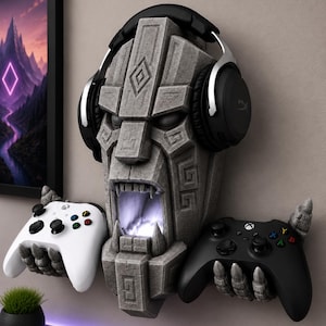 May include: A gray stone-textured wall mount shaped like a roaring face with two clawed hands extending to hold a white and a black gaming controller. The sculpture features a glowing light in the mouth and supports black over-ear gaming headphones.