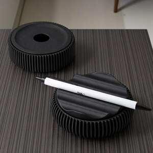 May include: Two black, textured desk organizers. One is a round container with a lid and a hole in the center. The other is a smaller, round organizer with a wavy top, holding a white and black pen with the word "BIC" printed on it.