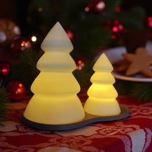 May include: Two glowing, frosted glass Christmas tree-shaped lights sit on a gray tray. The larger tree is to the left, and the smaller one is to the right. The lights are on a red tablecloth with a Christmas tree and ornaments in the background.