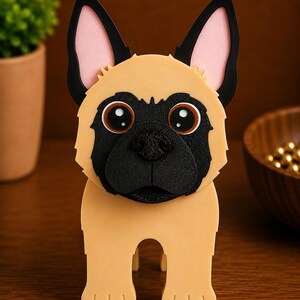 May include: A tan and black dog-shaped decorative item. The dog has large, round eyes, a black nose, and black ears with pink inner ears. The item is standing on a wooden surface.