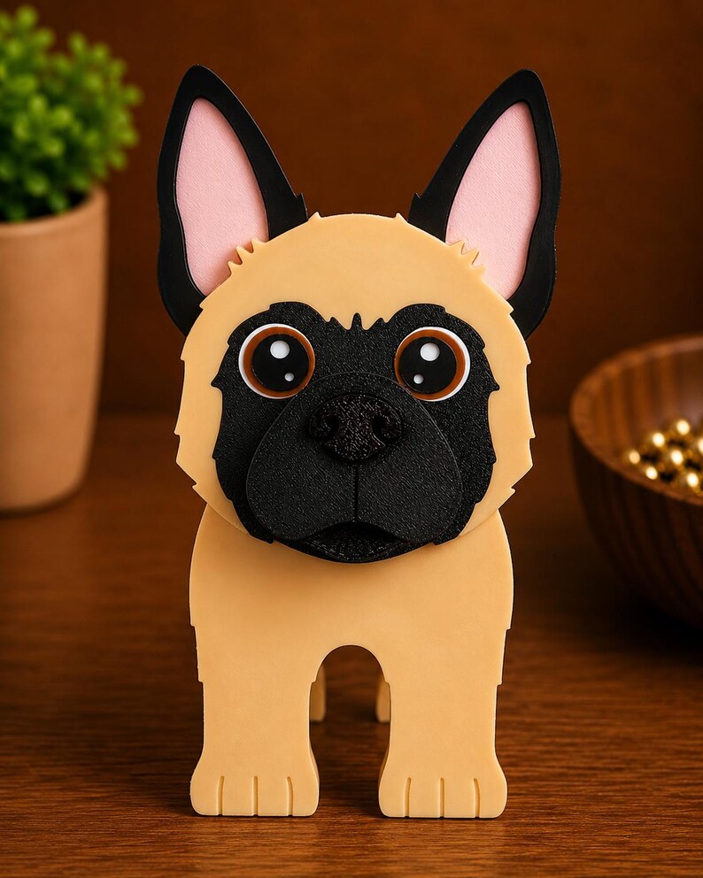 May include: A tan and black dog-shaped decorative item. The dog has large, round eyes, a black nose, and black ears with pink inner ears. The item is standing on a wooden surface.