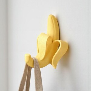 May include: A yellow banana-shaped wall hook, mounted on a white wall, holding a beige tote bag. The banana peel is partially open, revealing the banana inside. The hook is designed to resemble a peeled banana.