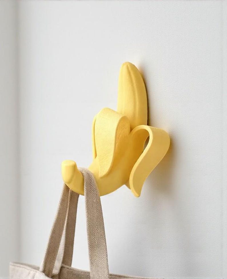 May include: A yellow banana-shaped wall hook, mounted on a white wall, holding a beige tote bag. The banana peel is partially open, revealing the banana inside. The hook is designed to resemble a peeled banana.
