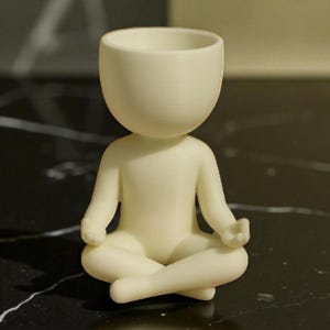 May include: A cream-colored figurine of a person in a seated yoga pose. The figure has a rounded head with a hollowed-out top, and the arms and legs are in a meditative position. The figurine is made of a smooth material.