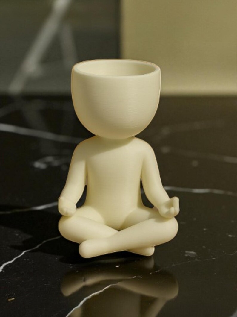 May include: A cream-colored figurine of a person in a seated yoga pose. The figure has a rounded head with a hollowed-out top, and the arms and legs are in a meditative position. The figurine is made of a smooth material.
