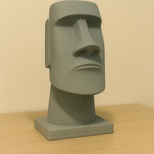 May include: A gray Moai statue replica, a stylized human head with a rectangular base. The sculpture features a prominent nose, defined lips, and large ears. The statue is displayed on a wooden surface.