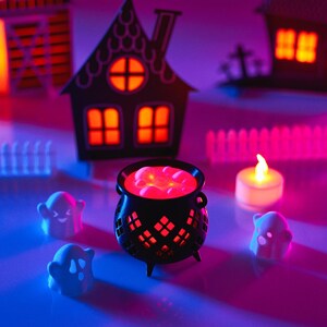 May include: A black cauldron with red glowing liquid inside, surrounded by three white ghost figurines and a small white candle. The scene is set on a table with a pink and blue glow, and there are paper houses in the background.
