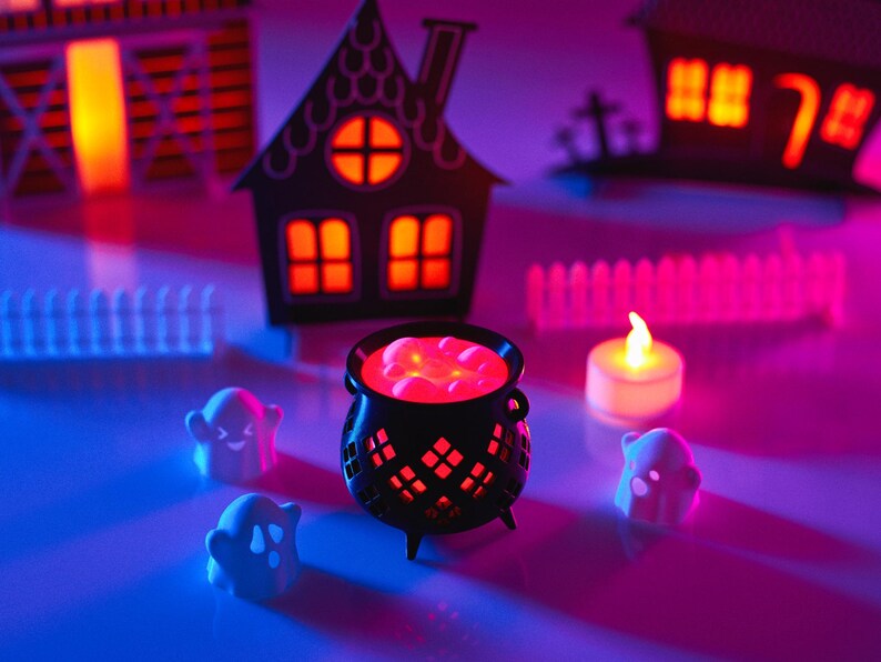May include: A black cauldron with red glowing liquid inside, surrounded by three white ghost figurines and a small white candle. The scene is set on a table with a pink and blue glow, and there are paper houses in the background.