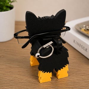 May include: A black and yellow dog-shaped eyeglasses holder. The holder has a black body with yellow paws and ears. The eyeglasses are held in place by the dog's head. A silver key ring is attached to the holder.