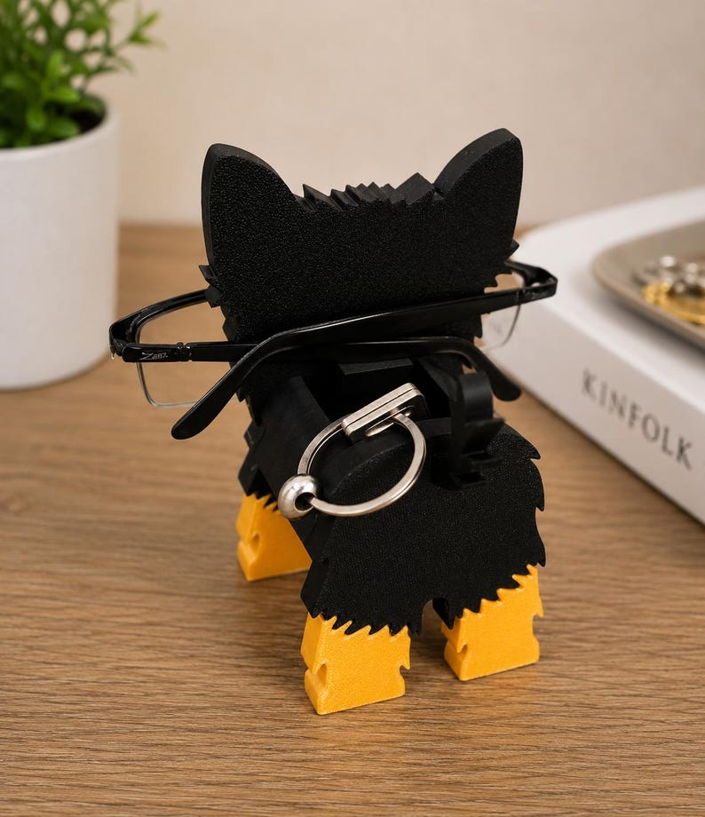 May include: A black and yellow dog-shaped eyeglasses holder. The holder has a black body with yellow paws and ears. The eyeglasses are held in place by the dog's head. A silver key ring is attached to the holder.