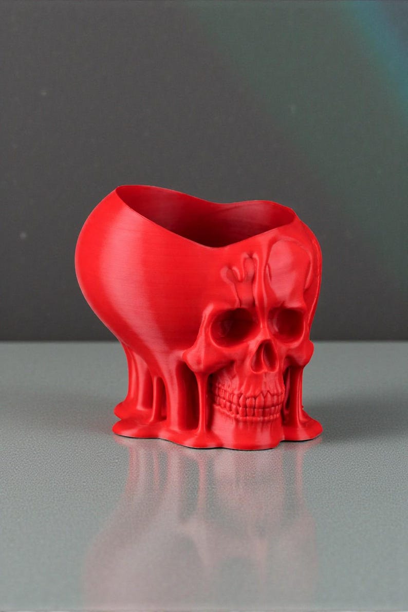 May include: A red skull-shaped planter with a heart-shaped opening. The planter has a textured surface and a detailed skull design, including eye sockets, a nose cavity, and teeth. The base has a melted appearance.