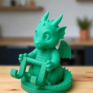 May include: A green dragon figurine holding a letter "J". The dragon has large eyes, small horns, and wings. The figurine is sitting on a round base and has a long tail.
