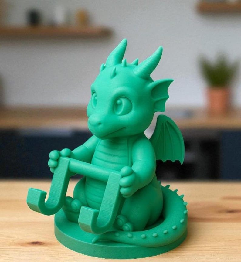 May include: A green dragon figurine holding a letter "J". The dragon has large eyes, small horns, and wings. The figurine is sitting on a round base and has a long tail.