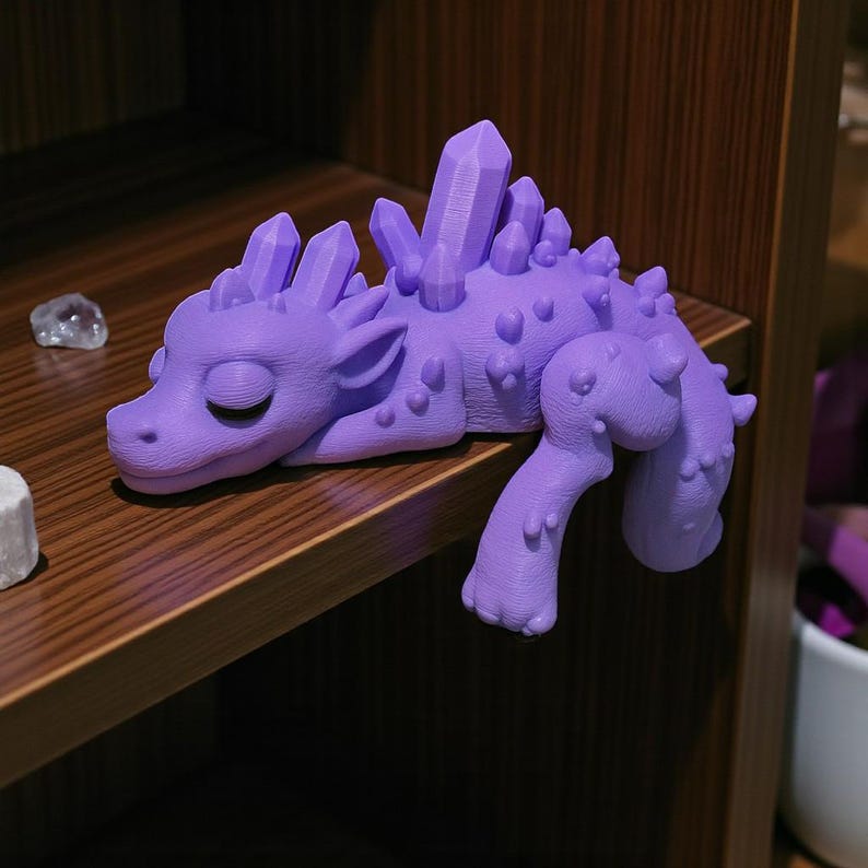 May include: A lavender-colored dragon figurine with crystal-like spikes along its back and head. The dragon is lying down, with its eyes closed. The figurine is resting on a wooden shelf.
