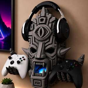 Könnte beinhalten: Ein grauer, texturierter Wandhalter in Form einer Steinmaske mit offenem Mund, der zwei klauenartige Hände besitzt, die einen weißen und einen schwarzen Gamecontroller halten, während ein schwarzer Over-Ear-Kopfhörer auf der Maske aufliegt.