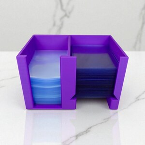 May include: A vibrant purple card holder with two compartments. One side holds a stack of blue-tinted card sleeves, the other a stack of dark blue sleeves. The holder is made of a solid material and sits on a white surface.