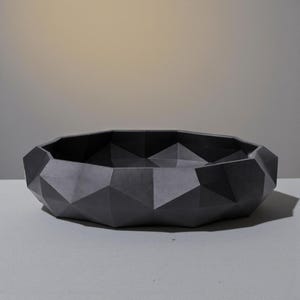 K&ouml;nnte beinhalten: Eine dunkelgraue, geometrische Schale mit facettiertem Design. Die Schale hat ein niedriges Profil und eine breite &Ouml;ffnung. Die Oberfl&auml;che ist matt und die Schale ist wahrscheinlich aus einem festen Material gefertigt. Geeignet f&uuml;r dekorative oder funktionale Zwecke.