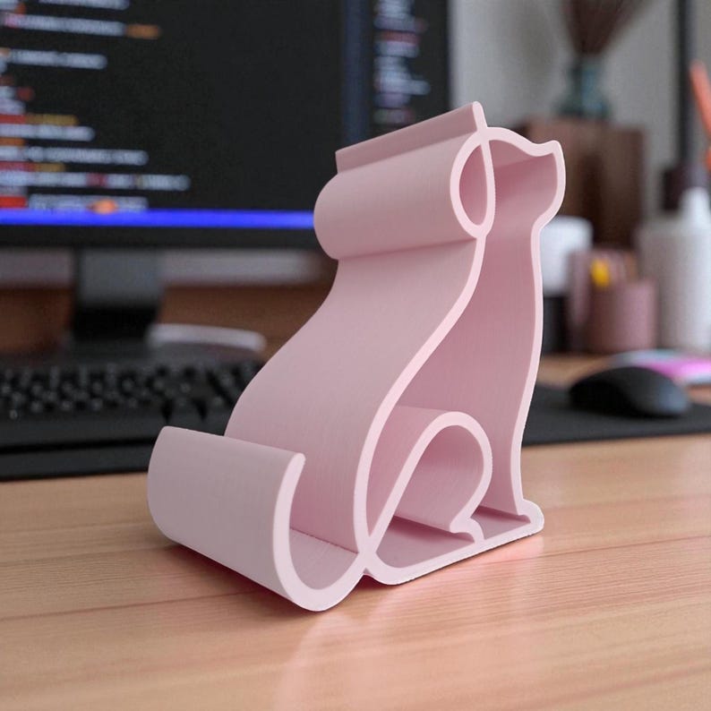 May include: A light pink phone stand shaped like a sitting dog. The stand is made of a solid material and has a curved design. The dog-shaped stand is on a wooden desk, with a computer and other office supplies in the background.