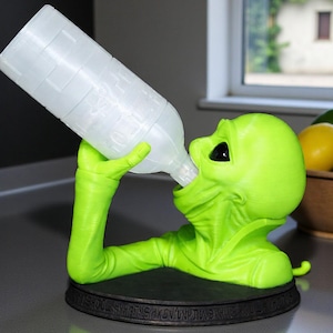 May include: A lime green alien figurine drinking from a clear plastic bottle. The alien is resting on a black circular base with etched text. The bottle is held in the alien's hands and tilted towards its mouth.