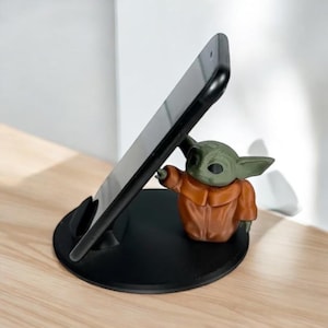 May include: Black phone stand with a green and brown Grogu figure. The phone stand is designed to hold a smartphone at an angle. The Grogu figure is pointing towards the phone. The stand is on a light brown surface.
