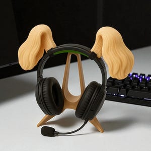 May include: Black headphones with a microphone rest on a wooden stand shaped like a dog's head, with tan-colored ear-shaped accents. The stand is on a white surface, with a black keyboard in the background.