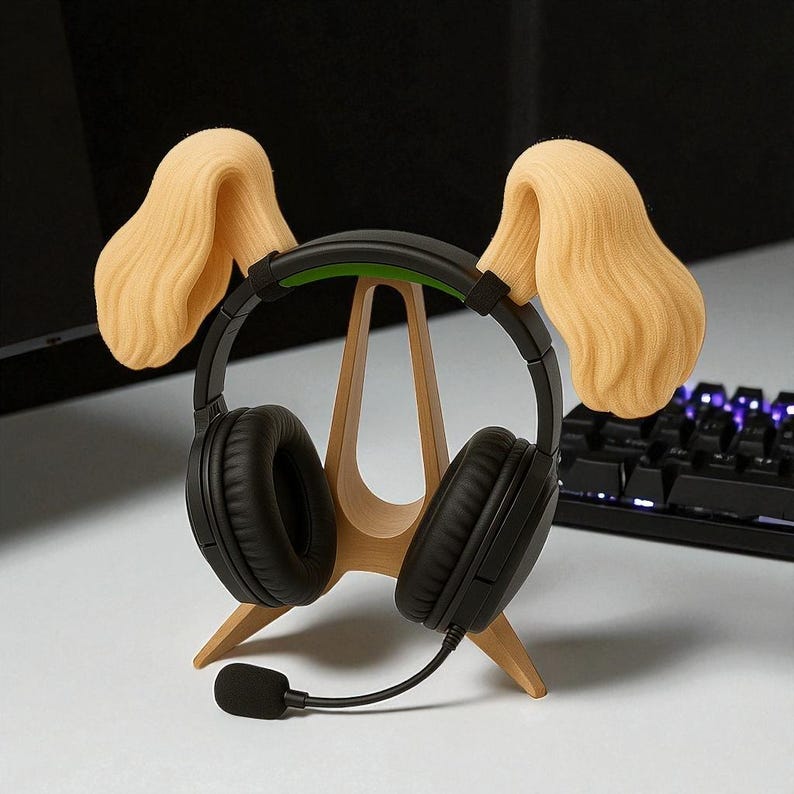 May include: Black headphones with a microphone rest on a wooden stand shaped like a dog's head, with tan-colored ear-shaped accents. The stand is on a white surface, with a black keyboard in the background.