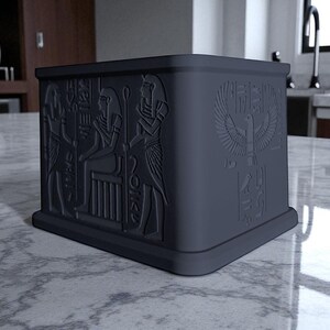 May include: A dark gray, square box with embossed Egyptian hieroglyphs and symbols. The box features detailed carvings of figures and an eagle with outstretched wings. The design is on all sides of the box.