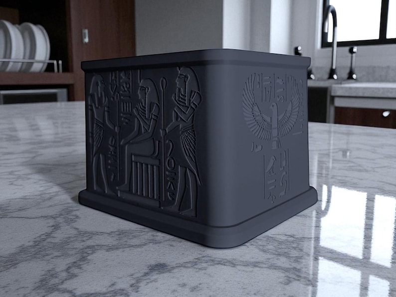 May include: A dark gray, square box with embossed Egyptian hieroglyphs and symbols. The box features detailed carvings of figures and an eagle with outstretched wings. The design is on all sides of the box.