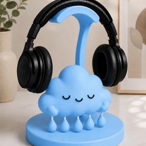 May include: A light blue headphone stand shaped like a cloud with a smiling face and rain drops. Black headphones rest on the stand. The stand is on a white surface.