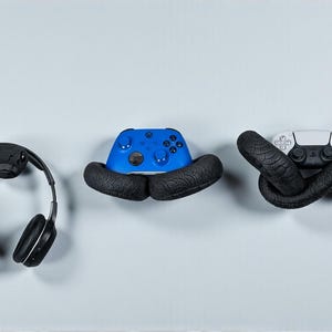 May include: Three gaming accessories: black headphones, a blue game controller, and a white game controller. Each item is held by a black snake-shaped holder. The items are displayed against a light blue background.