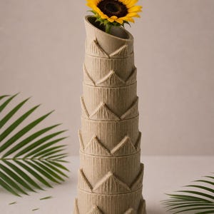 May include: A tall, beige vase with a textured, stacked design, holding a vibrant yellow sunflower. The vase has a unique, angled opening. The sunflower's dark center contrasts with its bright yellow petals.