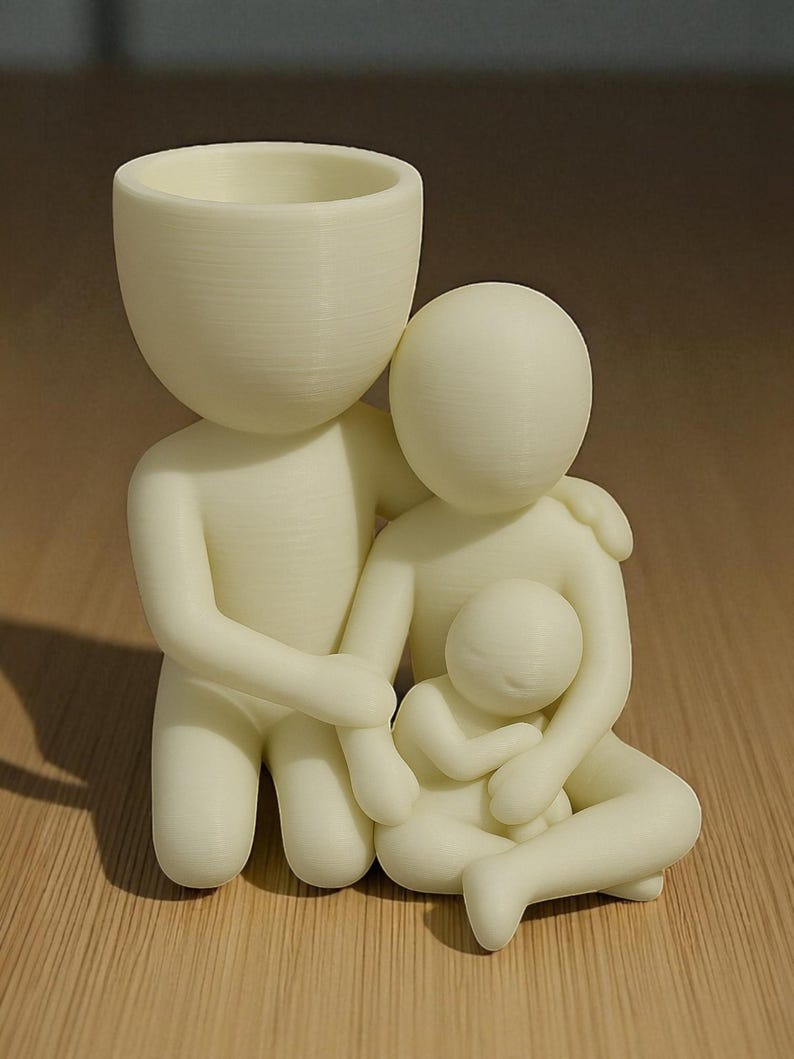 Puede incluir: Figurita familiar de color crema, que presenta una figura paterna con una cabeza grande y una peque&ntilde;a abertura para maceta, abrazando a otro padre y a un ni&ntilde;o. Las figuras est&aacute;n estilizadas y parecen impresas en 3D.