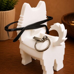 May include: A white dog-shaped holder for glasses and keys. Black-framed spectacles rest on the holder, and a set of keys is attached to the key ring. A small plant in a pot is in the background. The holder is designed to hold glasses and keys.