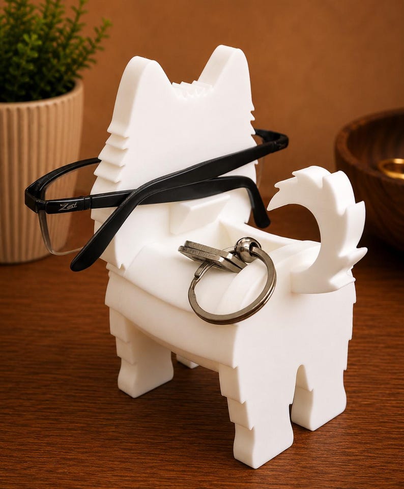 May include: A white dog-shaped holder for glasses and keys. Black-framed spectacles rest on the holder, and a set of keys is attached to the key ring. A small plant in a pot is in the background. The holder is designed to hold glasses and keys.