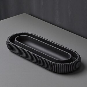 May include: A dark gray, oval-shaped decorative tray with a ribbed exterior and a smooth interior. The tray has a minimalist design and is suitable for various uses, such as holding jewelry or small items. The tray is approximately 12 inches long.
