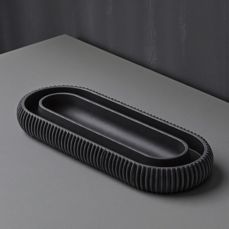 May include: A dark gray, oval-shaped decorative tray with a ribbed exterior and a smooth interior. The tray has a minimalist design and is suitable for various uses, such as holding jewelry or small items. The tray is approximately 12 inches long.