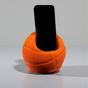 May include: An orange basketball-shaped phone holder with a black smartphone inside. The holder has the typical basketball texture and lines. The phone is upright, filling the holder's opening. The background is a plain, light grey.