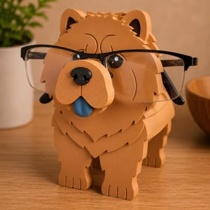 May include: A tan-colored Chow Chow dog-shaped eyeglasses holder with black-framed glasses. The holder has a blue tongue and black nose. The holder is made of a solid material and sits on a wooden surface.