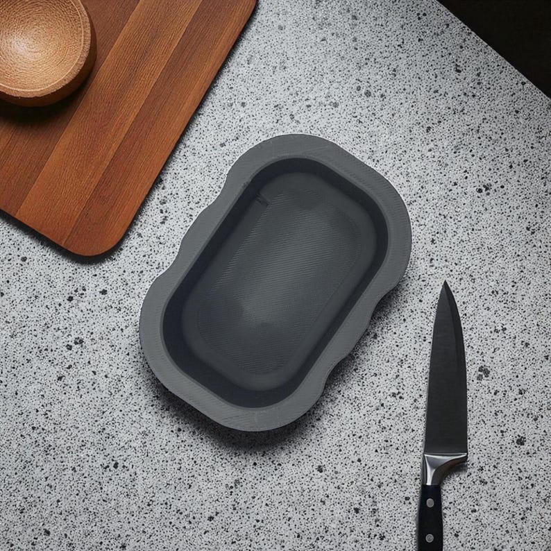 May include: Overhead shot of a gray silicone dish, a wooden cutting board, a small wooden bowl, and a black chef's knife on a speckled gray countertop. The dish has a rectangular shape with rounded edges.