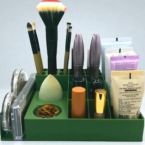 May include: A green makeup organizer filled with various cosmetic items. It contains makeup brushes, mascara, lipsticks, a makeup sponge, and other beauty products. The organizer is designed to keep beauty supplies neat and accessible.
