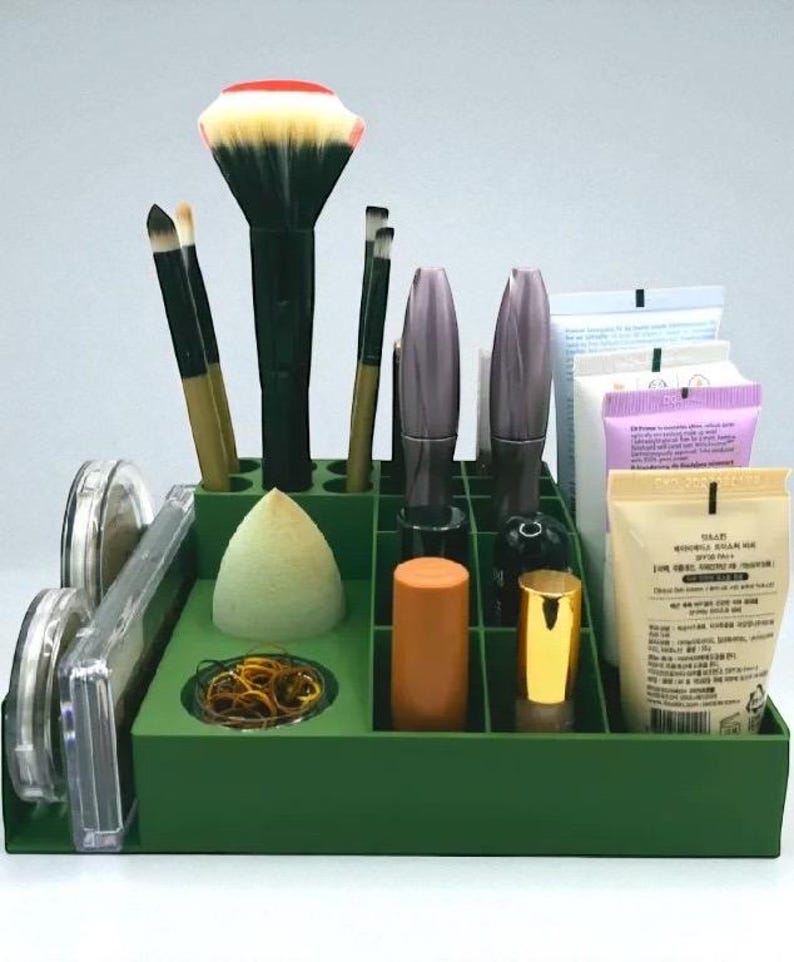 May include: A green makeup organizer filled with various cosmetic items. It contains makeup brushes, mascara, lipsticks, a makeup sponge, and other beauty products. The organizer is designed to keep beauty supplies neat and accessible.